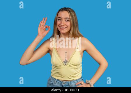 Giovane donna felice che mostra un segno ok con la mano e sorride su sfondo blu Foto Stock