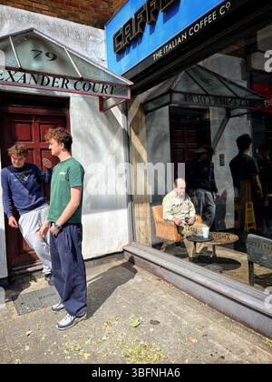 Due giovani, che sembrano gemelli, stanno fuori da un caffè, mentre all'interno del bar, un uomo calvo anziano si siede a un tavolo da solo. Foto Stock
