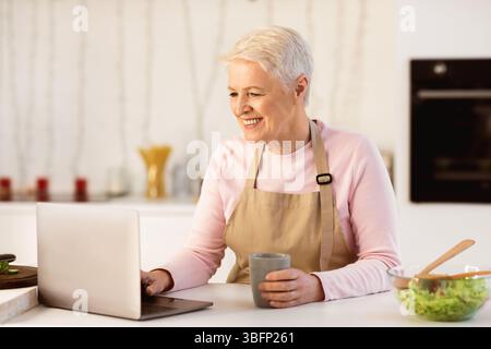 Donna anziano felice usando il laptop che guarda video di cottura in cucina Foto Stock