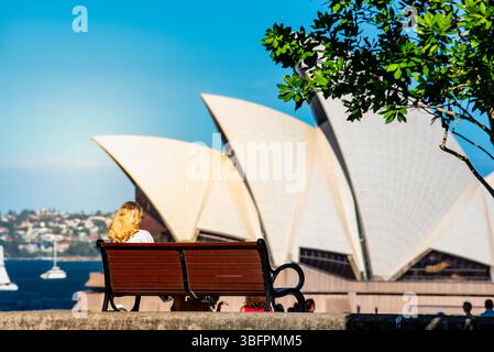 Una giovane donna bionda siede su una panchina del parco guardando dall'altra parte della Sydney Opera House in una giornata di sole in Australia Foto Stock