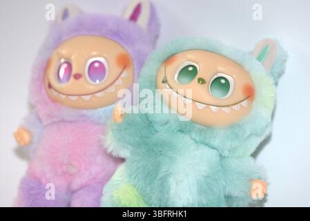 Due giocose figure di giocattoli Labubu in morbidi costumi da mostro verde e rosa e un sorriso malizioso, posate su uno sfondo bianco. Foto Stock