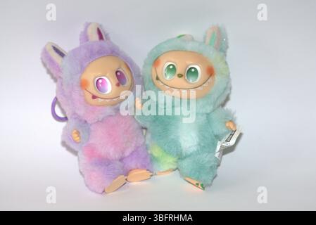 Due giocose figure di giocattoli Labubu in morbidi costumi da mostro verde e rosa e un sorriso malizioso, posate su uno sfondo bianco. Foto Stock