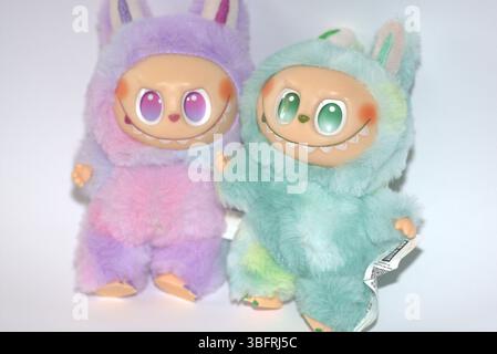 Due giocose figure di giocattoli Labubu in morbidi costumi da mostro verde e rosa e un sorriso malizioso, posate su uno sfondo bianco. Foto Stock