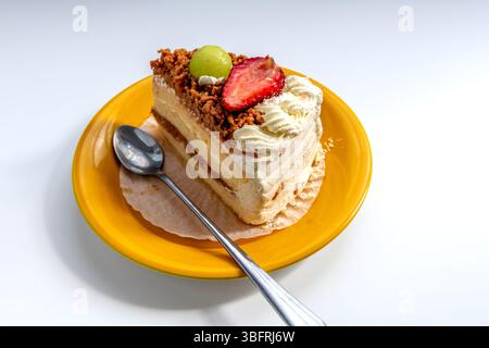 Una fetta di torta alla panna condita con fragole e uva verde servita su un piatto giallo isolato su bianco. Foto Stock
