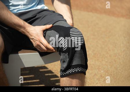 Uomo seduto su una panchina che indossa un ginocchio per sostenere una gamba dolorosa. Gamba dell'atleta che indossa un sostegno per le ginocchia o un bendaggio elastico per le ginocchia. Foto Stock