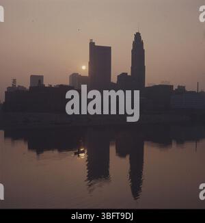 Una serena vista mattutina dello skyline di Columbus visto dal fiume Scioto, con una canoa che scivola sull'acqua. La luce del mattino illumina gli edifici del centro, mostrando la vibrante atmosfera della città. Foto Stock