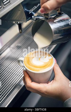 Il barista prepara un cappuccino, versando la schiuma di latte in una tazza di caffè creando una latte art Foto Stock