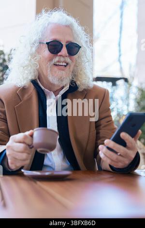 Ritratto verticale di un anziano con un caffè che usa lo smartphone in un bar all'aperto Foto Stock