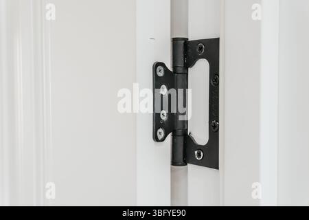 Primo piano di una robusta cerniera in metallo nero fissata saldamente a un moderno telaio bianco della porta, evidenziando i dettagli del meccanismo e delle viti Foto Stock