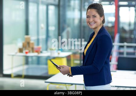 Donna che tiene un tablet con stilo e indossa un blazer blu navy in un ufficio aperto con prototipi e spazio di copia Foto Stock