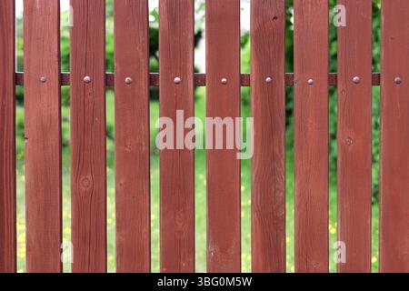 Primo piano della recinzione in tavole di legno dipinte di rosso con un giardino verde sfondo sfocato Foto Stock