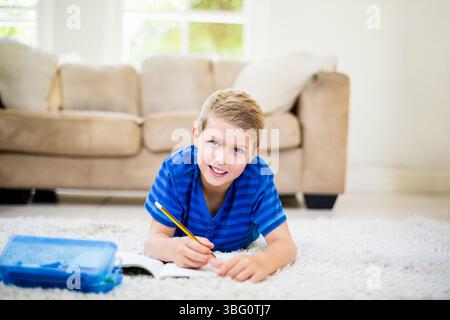 Ragazzo maschio in età scolare sdraiato su un tappeto di peluche nel soggiorno disegnando su un notebook con matita gialla Foto Stock