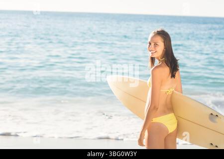 Surfista che tiene in mano la tavola da surf, guarda indietro verso la macchina fotografica sulla spiaggia in costume da bagno giallo, copia spazio Foto Stock