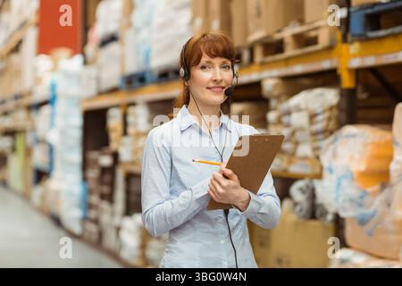 Donna responsabile dell'inventario che ispeziona le scorte nella corsia del magazzino con portadocumenti a matita e cuffie Foto Stock