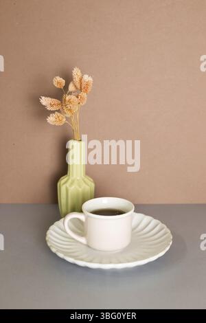 Tazza di tè e vaso di pianta secca rosa su tavola grigia. Parete di fondo marrone Foto Stock