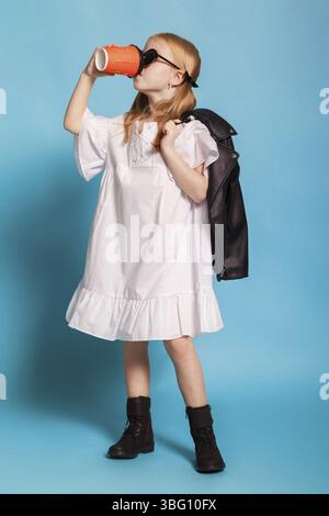 Isolata su una ragazza blu dai capelli rossi con code in occhiali da sole, abito bianco e scarpe stringate, con una giacca di pelle nera e bevendo da un arancio Foto Stock