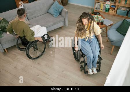 Giovane uomo caucasico con disabilità manovrando sedie a rotelle con amici femminili mentre si diverte in salotto, festeggiando il compleanno insieme, gioco da tavolo visibile sul tavolo Foto Stock