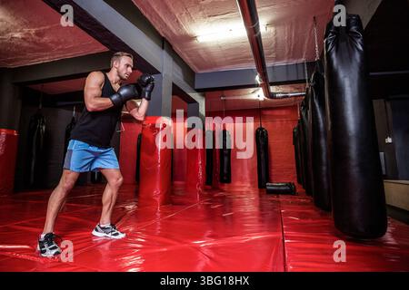 Uomo che pratica pugilato nel seminterrato della palestra, con un sacco pesante con guanti su tappetini rossi, spazio per copiare Foto Stock