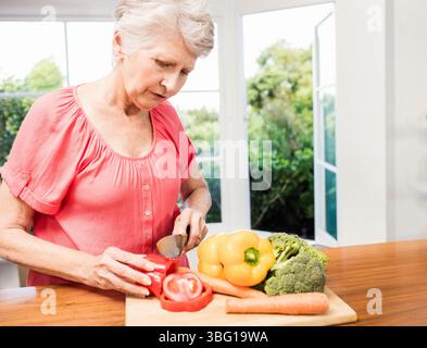 Donna anziana che indossa una camicetta di corallo che affetta peperone rosso sul tagliere in cucina, spazio copia Foto Stock