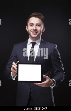 Giovane uomo d'affari di successo in un elegante abito da lavoro su sfondo nero, sicuro di tenere il tablet davanti e sorridente Foto Stock