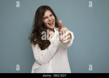 Ritratto di giovane positivo felice allegro attraente donna ricci bruna con emozioni sincere indossando casual camicia bianca isolato su sfondo blu w Foto Stock