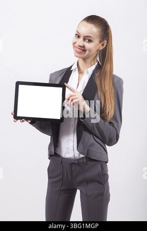 Giovane donna d'affari moderna con un costume da lavoro su sfondo bianco che tiene in avanti lo schermo del tablet e sorride con i denti bianchi Foto Stock
