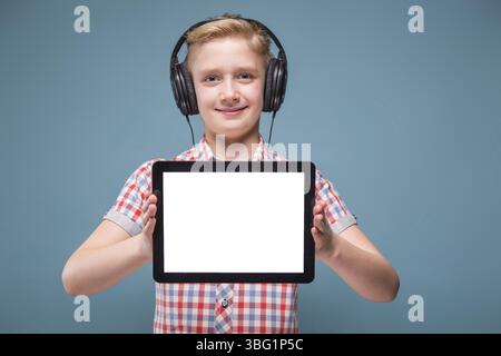 Studente biondo sorridente con le cuffie che tengono il telefono a distanza di braccia e mostra il display, lo studio lap ha girato una foto di adolescenti europei con il reparto Foto Stock