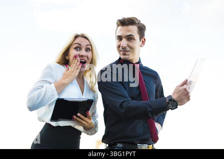 Coppia di uomini d'affari, giovane bruna dall'aspetto carino uomo caucasico con vestito scuro, cravatta rossa e camicia nera, e attraente donna bionda in camicetta bianca Foto Stock