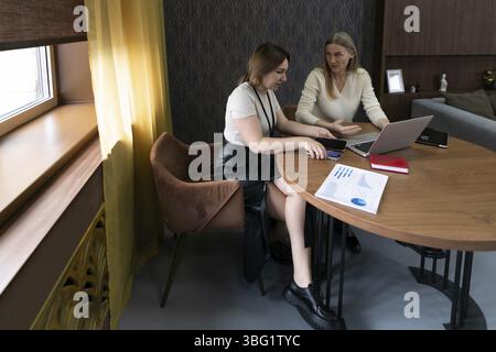 In un ufficio elegante, due donne si impegnano in una conversazione mirata mentre esaminano i documenti su un notebook. La luce solare filtra attraverso la finestra, creando un Foto Stock