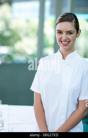 Donna che indossa un'uniforme bianca sorridente in una camera spa vicino a una grande finestra con asciugamani, occhiali, spazio per fotocopie Foto Stock