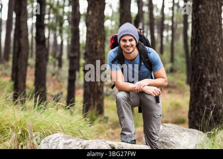 Uomo che si inginocchia su un grande masso nella pineta, indossa berretto e stivali da trekking con zaino rosso Foto Stock