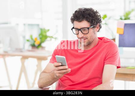 Uomo di mezza età che indossa una camicia rossa seduto alla scrivania tenendo lo smartphone tra i monitor del computer Foto Stock