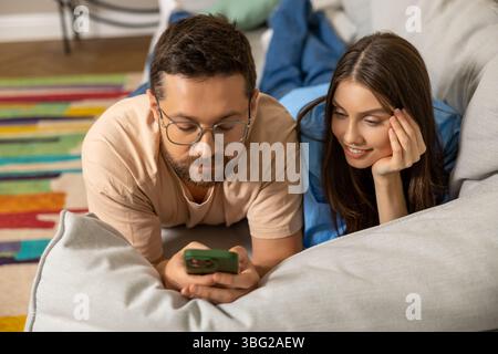 Giovane donna felice e uomo che usano lo smartphone insieme sdraiati sul divano Foto Stock