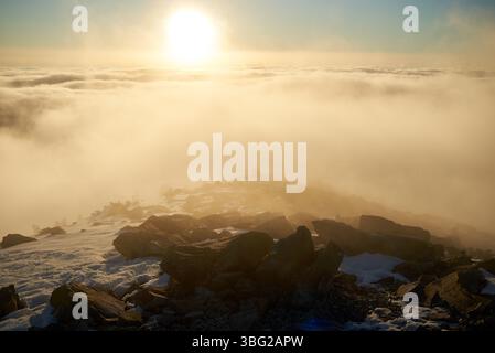 Alba sulla cima innevata della montagna. La calda luce dorata del sole che sorge bagna le nuvole in delicate sfumature di arancione e rosa. Neve e rocce sparse in primo piano aggiungono texture e profondità alla scena. Foto Stock