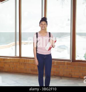 Studentessa adolescente sorridente che tiene libri di testo e zaino nel corridoio della scuola con finestre innevate Foto Stock