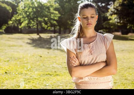 Donna in piedi con le braccia incrociate che guardano verso il basso in un prato illuminato dal sole, indossa un abito rosa pallido, copia spazio Foto Stock
