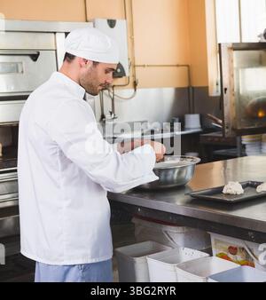 Uno chef maschile di 30 anni indossa un cappotto da chef che forma l'impasto in cucina commerciale con una ciotola per mescolare Foto Stock