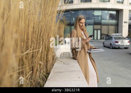 Una donna d'affari con lunghi capelli biondi indossa un cappotto beige. Sta fuori da un business center e sta usando il telefono. Ha un taccuino dentro di lui Foto Stock