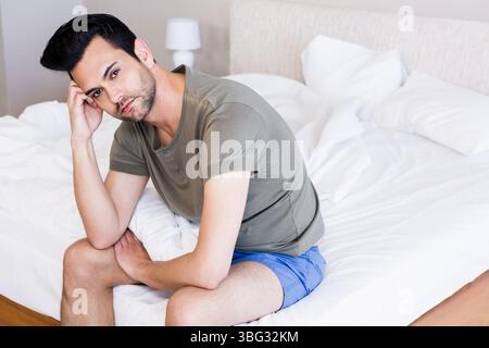 Uomo medio adulto seduto sul bordo di un letto non fatto in camera da letto con lampada per comodino che tiene la testa Foto Stock