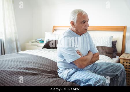 Uomo dai capelli bianchi seduto sul letto vicino al comodino con libri, cuscini, che guarda direttamente in camera da letto Foto Stock