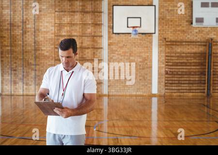 Allenatore adulto maturo che tiene in mano appunti con penna vicino al canestro da basket e ai bar a parete in palestra Foto Stock