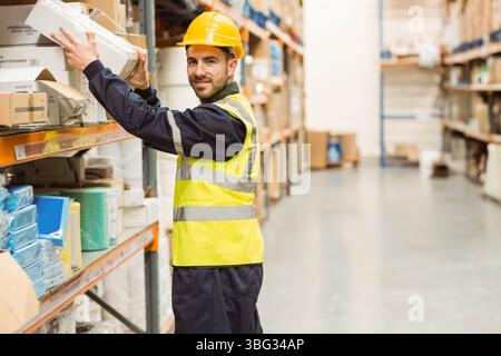 Lavoratore di magazzino che indossa elmetto, giubbotto che raggiunge la scatola su scaffale metallico nella corsia del magazzino Foto Stock