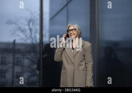 Una donna anziana alla moda, vestita elegantemente con un cappotto leggero, sorride mentre parla al telefono fuori di un edificio di vetro contemporaneo, sentendosi confidente Foto Stock
