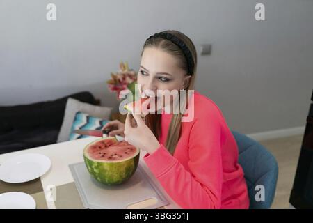 Una ragazza che indossa un abito luminoso prende un boccone di anguria mentre si siede a un tavolo da pranzo. L'ambiente vivace della cucina aggiunge un'atmosfera rinfrescante Foto Stock