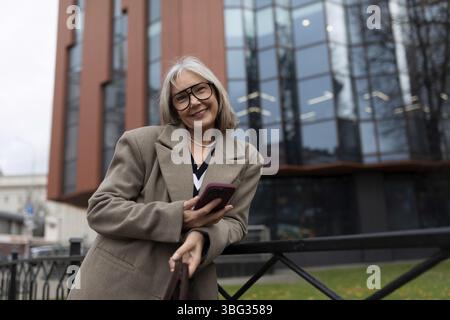 La donna d'affari anziana si posa all'esterno di un moderno edificio di uffici, vestito con un cappotto elegante. Sorride e tiene il suo smartphone, trasuda confidenza Foto Stock