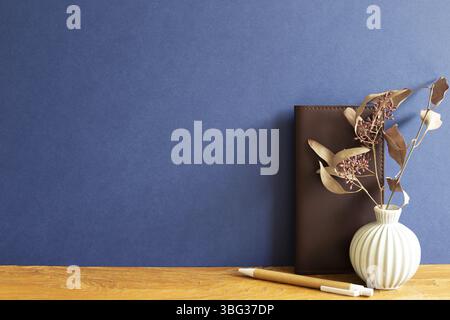 Notebook in pelle marrone, penna, fiori secchi su scrivania in legno. Sfondo blu Foto Stock