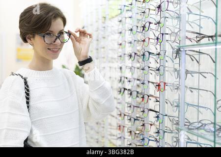 Foto di giovane donna bruna pensante attraente in un maglione bianco sceglie gli occhiali per la visione su una vetrina del negozio Foto Stock