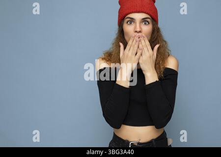 Scatto di giovane emotivo stupito bella bruna donna winsome capelli ondulati con emozioni sincere indossando nero crop top e cappello rosso isolato su blu bac Foto Stock