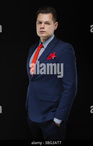 Giovane uomo d'affari caucasico elegante e intelligente in tuta blu in piedi in ufficio con le braccia in tasca Foto Stock