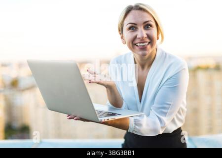 La giovane, bella e seria donna d'affari caucasica con camicetta bianca e gonna nera si staglia sul tetto con un laptop argentato in mano, esaltante, stupita Foto Stock